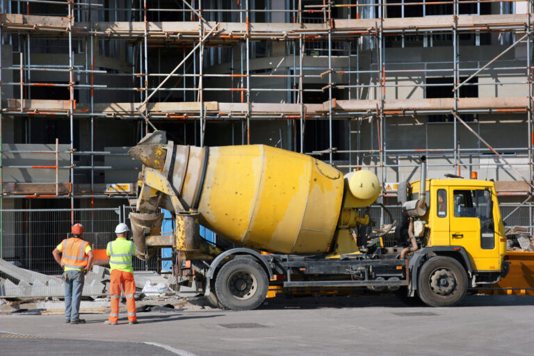 Yellow cement mixer truck parked in fornt of a new building under construction with scaffolding in position. Two workmen standing to the left hand side, rear view.